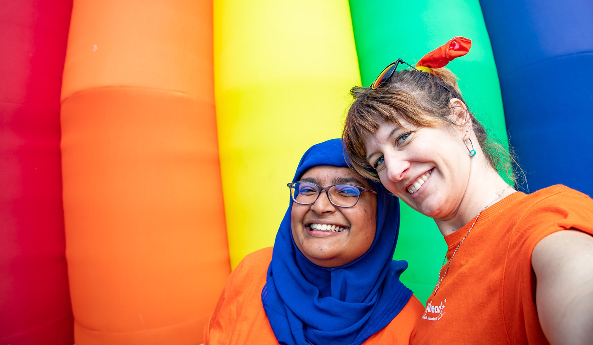 Two smiling WayAhead staff in front of a rainbow background from Fair Day