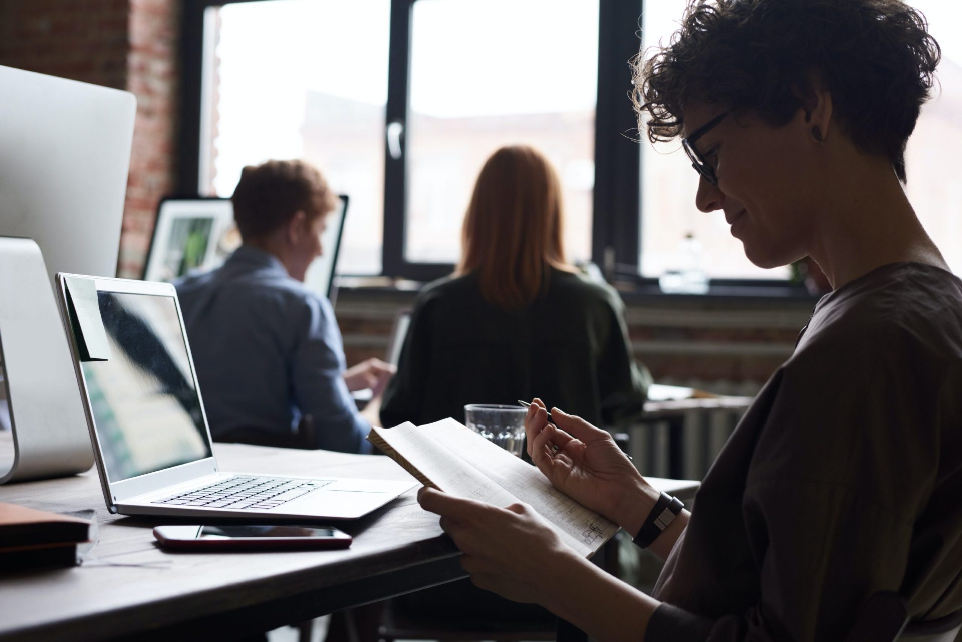 Woman at work writing in note book, slightly in silhouette with other people working behind